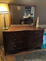Photo of wide wooden chest of drawers with dark wood finish and attached rectangular mirror on back. Shows lamp and wall clock reflections.