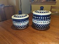 Two Polish pottery jars with lids on a wooden table, showing decorative patterns and size difference.