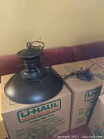 Image showing the black metal hanging light fixture resting on cardboard boxes, displaying the exterior side and hanging chain.