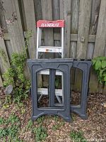 Featherlite aluminum 3 step ladder with red top and two black plastic work horses placed against a wooden fence outdoors on ground with leaves.