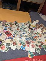 Large number of assorted worldwide stamps scattered on a bed near a large manila envelope.