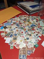 A large assortment of loose stamps spread on a red surface with an envelope and books in the background.