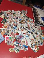 Loose assorted stamps scattered on a red surface showing variety in size, color, and design from multiple countries.