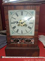 Front view of wooden mantel clock with Roman numeral dial and glass door closed, showing decorative lower panel and measuring tape for scale.