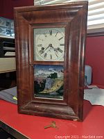 Front view of the wooden mantel clock showing the clock face with Roman numerals and the reverse painted lower glass panel with a scenic landscape.