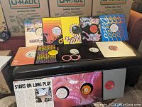 Wide shot of 14 disco vinyl records displayed on a black bench, showing covers and vinyl labels, with U-Haul boxes in background.