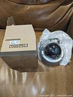 Photo showing the dome-style black-tinted vandal-proof security camera with attached power cable and protective soft packaging, placed next to its cardboard box.