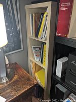 Side angle photo showing full height of beige/light wood IKEA bookcase with four compartments filled with books and papers, next to a wooden side table with lamp.