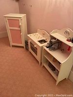 Wide view of the three-piece wooden play kitchen set placed against a wall with pink paint. Includes tall cabinet, stove/sink unit, and hutch.