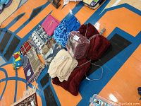 Lot of 11 blankets displayed on floor: 4 burgundy Sunbeam heated throws (one sealed in plastic), a Wonder Woman sleeved blanket, beige fleece throw, and 5 assorted tasseled throw blankets including plaid and patterned designs. Various colors and patterns visible.