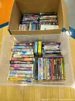 Top down photo showing two boxes, one cardboard one plastic crate, filled with various VHS and DVD movie cases, mostly children and animation titles visible on spines, stacked tightly.