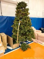 Full view of 9 ft pre-lit Christmas tree with clear lights, standing assembled, with folded smaller tree pieces on floor and boxes nearby.