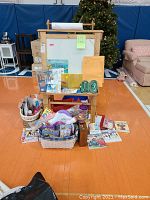 Front view of double-sided wooden easel with paper dispenser, surrounded by multiple craft supplies baskets, art books, and kits placed around the base on gym floor.