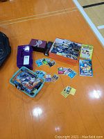 Wide view of the lot showing Pokemon trading cards spread out, a box of additional cards, Pacific Giftware chalice, Avengers lunchbox, and plastic storage containers.