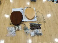 Two wooden elongated toilet seats (dark and light wood) with boxes underneath, chrome shower organizer, assorted hardware pieces in bags and loose on floor.