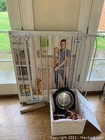 Photo of white metal adjustable entryway gate in original box and fabric storage box containing pet supplies, placed in front of glass door.