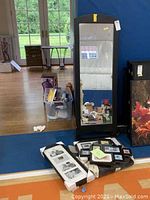 Photo showing tall free standing black framed floor mirror with visible wear on edges, positioned next to plastic bin of miscellaneous items and folded frames.
