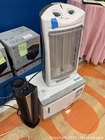 Sunbeam towel warmer heater in white rectangular design with control dials and metal grill sits on a box along with a black and white LifeZone heater box nearby, a black tower heater on the floor