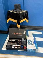 Magician's table with black cloth and gold trim, black magician's hat on top, magic set case on floor with various props inside, and Mind Flex game box next to it.