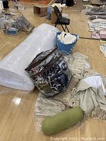 King mattress topper rolled and wrapped in plastic lying on floor with assorted blankets and bedding items around it.