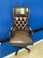 Front view of the brown leather office chair showing tufted backrest, nailhead trim, damaged sections on arms, and caster wheels.
