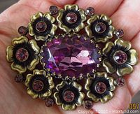 Front view showing the large oval facetted purple glass stone and surrounding floral brass-tone border with smaller purple stones.