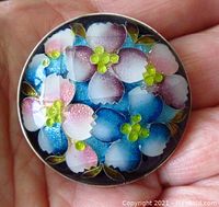 Front view of the brooch showing colorful enamel flowers in pastel pink, blue, and yellow on a round silver base held in hand.