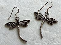 Front view of two sterling silver dragonfly earrings displaying detailed wing veins and segmented body design, silver color, hanging hook wires.