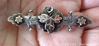 Front view of the antique sterling silver bar brooch showing gold floral decorations, engraved details and beaded edge, held in hand for scale.