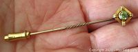 Close-up view of the stick pin held in hand showing twisted design, gold marking, and green peridot stone.