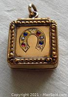 Front view of the small square gold locket with an engraved horseshoe set with small stones including seed pearls, rubies, and sapphires.