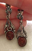 Front view of pair of sterling silver earrings with cognac amber oval cabochons and detailed floral leaf designs held in hand.