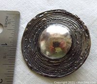 Top-down view showing the circular design with concentric lines and central dome, alongside a ruler for scale approximately 2 inches in diameter.