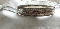 Front view of the sterling silver floral engraved bangle laying on fabric, showing the detailed engraving pattern and safety chain.