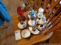 Overview of 7 assorted miniature oil lamps of various designs on wooden chair, mostly porcelain with floral designs, some with glass chimneys, some missing chimneys.