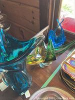 Three chalet style art glass pieces on a wooden surface near window, showing their translucent colors and organic shapes.