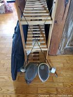 Two fencing masks with mesh front, two fencing foils with round guards and handles, and a black bag behind them, all leaning against a wooden rack.