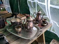 Photo showing two silver plated tea and coffee service sets on a large ornate tray, including pots, creamers, sugar bowls