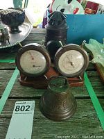 Photo showing miner's lamp, dual barometer set, and cast iron bell together on a wooden surface.