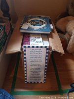 Box with assorted books including Chicken Soup for the Soul and Atlas of Beer, plus visible part of wall hanging with grandparent poem