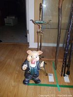 Photo of pig figurine and brass ashtray stand on wooden floor near glass cabinet.