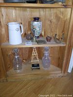 Wide view showing shelf with white Wedgwood pitcher, Chinese vase, art glass paperweight, brass bell, horseshoe, wooden box, glass decanters, and other small items.