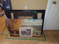 Four artworks stacked against wall: original landscape oil painting showing autumn trees and river, vintage lithograph partially covering painting, wooden frames behind them.