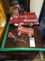Overview of all three vintage toys on a dark wooden surface showing the red Tonka fire truck, red wooden dog toy with wheels, and a cast iron horse-pulled fire truck figurine with firemen figures.