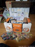 White/orange cardboard crate holding assorted loose Pokemon cards spread on floor, folded playmat visible.