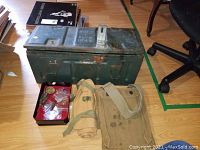 Photo showing green metal ammunition box, folded canvas bags, and a red tray with bullet shells and pins on the floor near chair