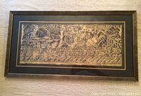 Framed artwork showing a horizontal battle scene in gold on black paper, featuring lines of soldiers, trees, and an elephant with combatants.