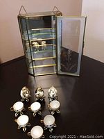 Etched glass display case open with miniature eggshell teacups and creamers placed outside on the table