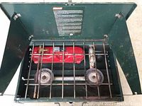 Top-down view of green Coleman 425 camping stove in metal case with red fuel bottle inside