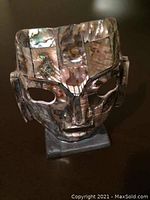 Front and angled views of a Mexican Aztec warrior mask made of abalone shell and obsidian stone on a stand, approximately 5 inches tall.